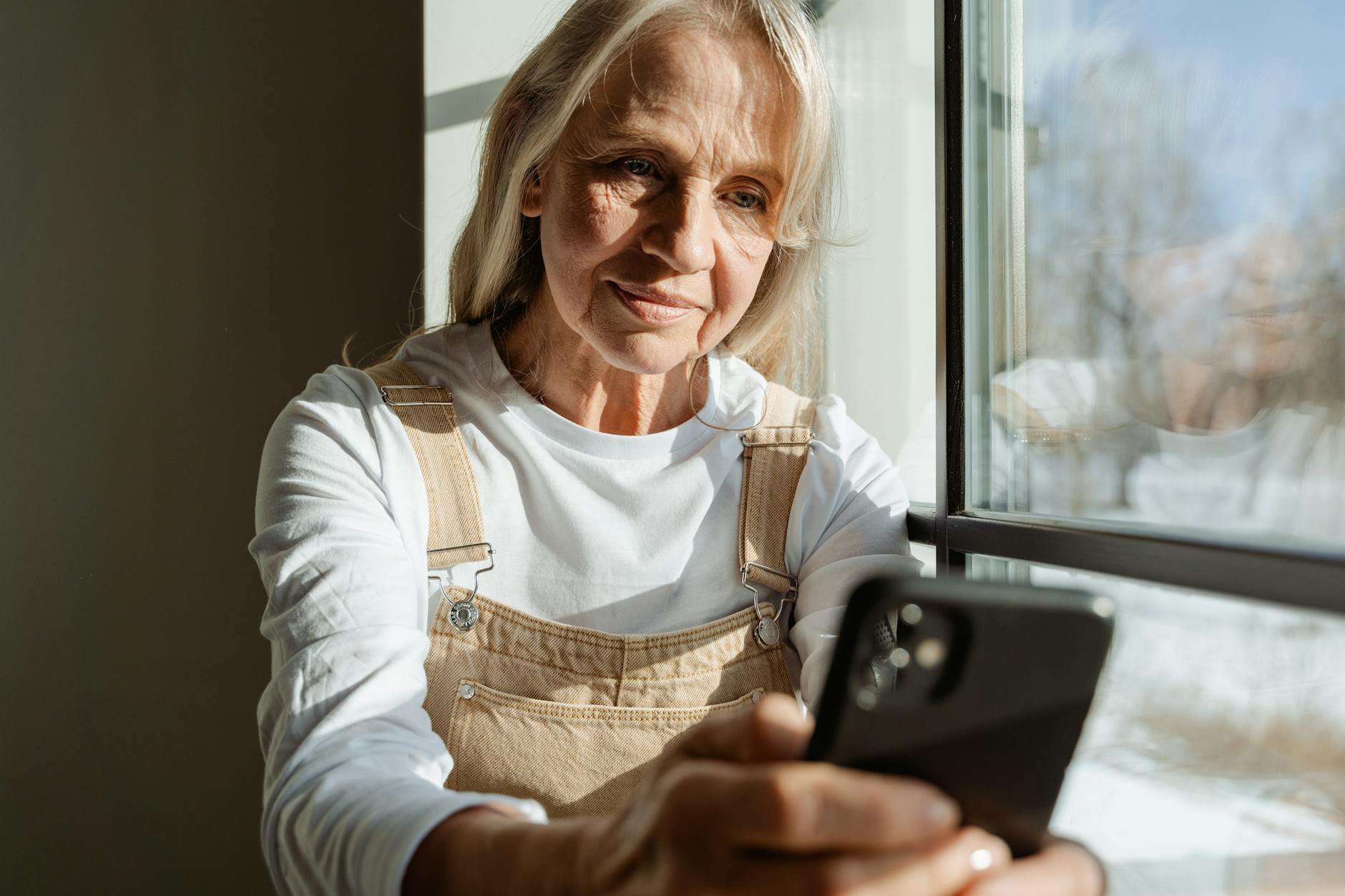 A senior woman enjoys leisure time browsing on her smartphone by a sunlit window.