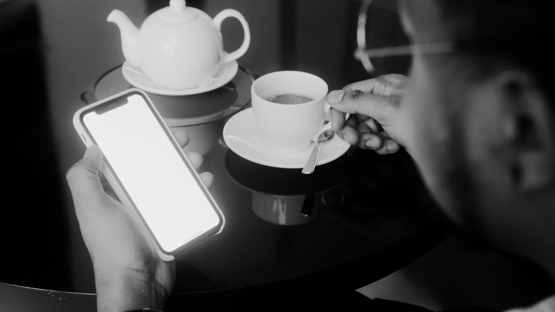 A grayscale close-up of a man holding a smartphone and a cup of tea.