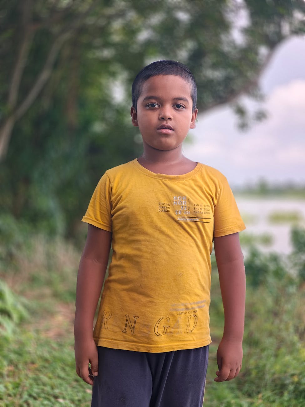 Portrait of a young boy in a yellow shirt standing outdoors with green nature background.