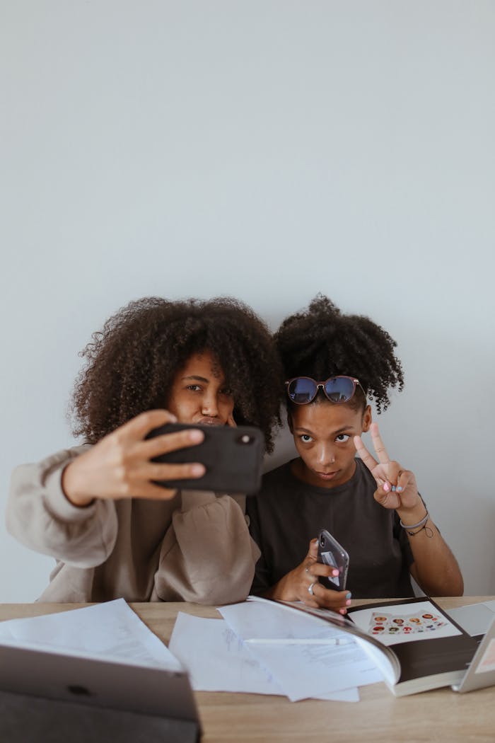 our-story-02 Two friends with afro hair taking a selfie indoors with smartphones and gadgets, creating joyful memories together.