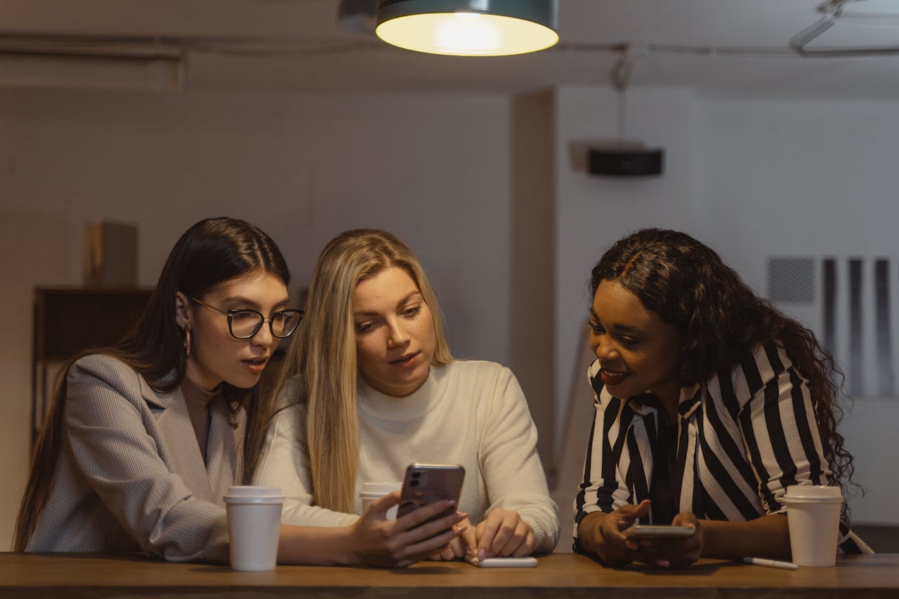 our-story-01 Three diverse women enjoying time together, interacting with smartphones at a table indoors.
